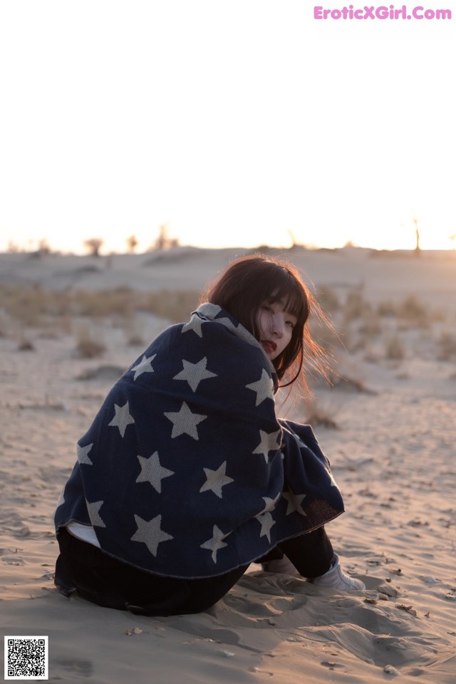 A woman wrapped in a blanket sitting in the sand.